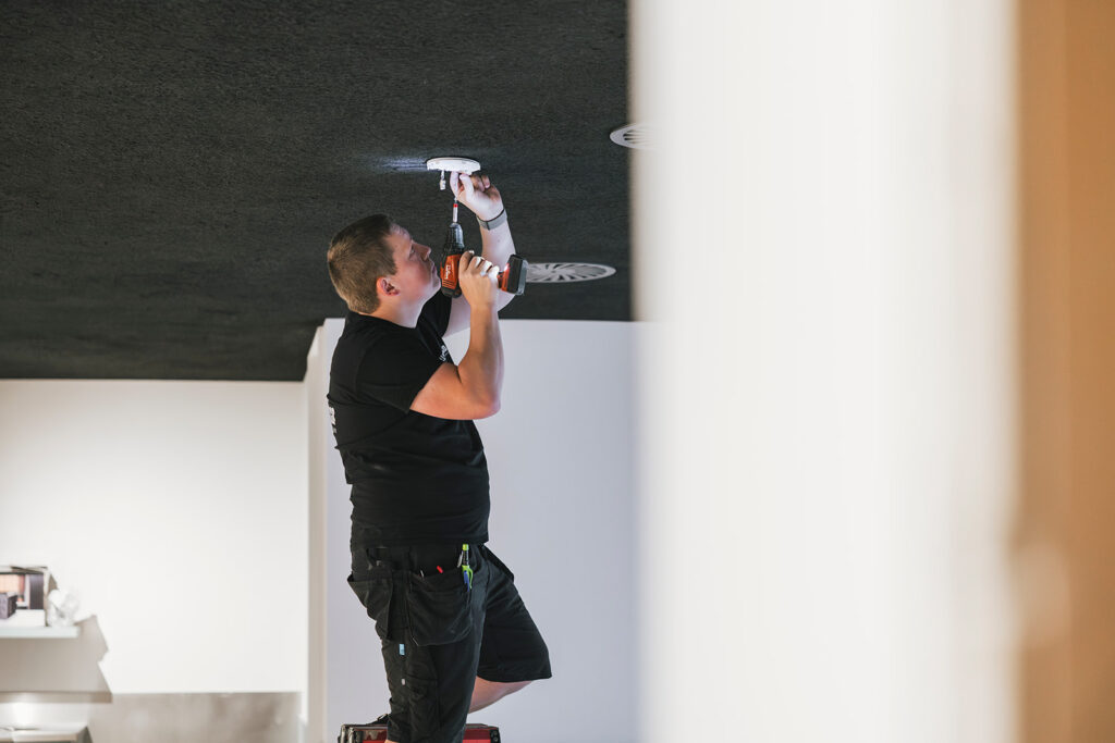 elektricien installeert rookmelder aan zwart plafond met accuboormachine in modern interieur